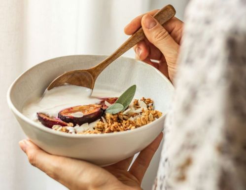Personne tenant un bol de yaourt garni de fruits, granola et une feuille de sauge, en utilisant une cuillère en bois.