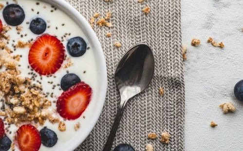 Bol de yaourt garni de tranches de fraises, myrtilles et granola, sur une serviette grise avec une cuillère en métal à côté.